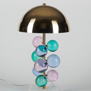 May include: A decorative table lamp with a gold-colored dome shade and a brass stem. The stem supports a cluster of colorful glass spheres in shades of pink, blue, and green. The lamp sits on a white marble base.