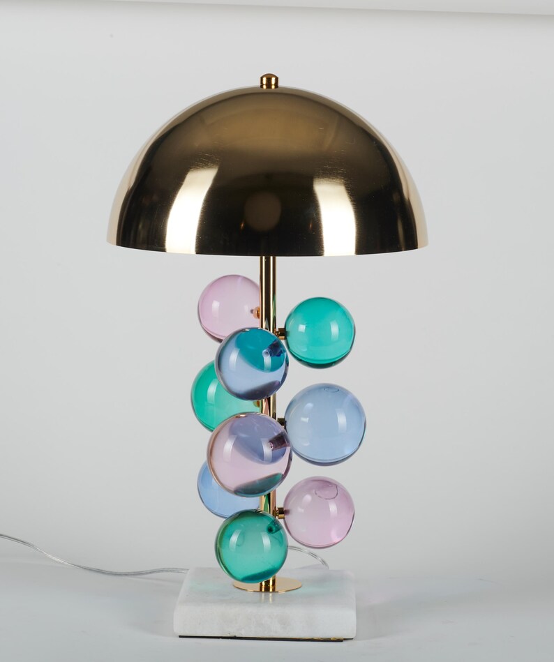 May include: A decorative table lamp with a gold-colored dome shade and a brass stem. The stem supports a cluster of colorful glass spheres in shades of pink, blue, and green. The lamp sits on a white marble base.