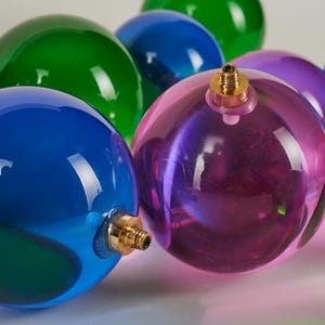 May include: A collection of colorful, transparent glass ornaments in shades of blue, green, and purple. Each sphere has a gold-colored metal cap. The ornaments are arranged closely together, reflecting light and creating a festive display.
