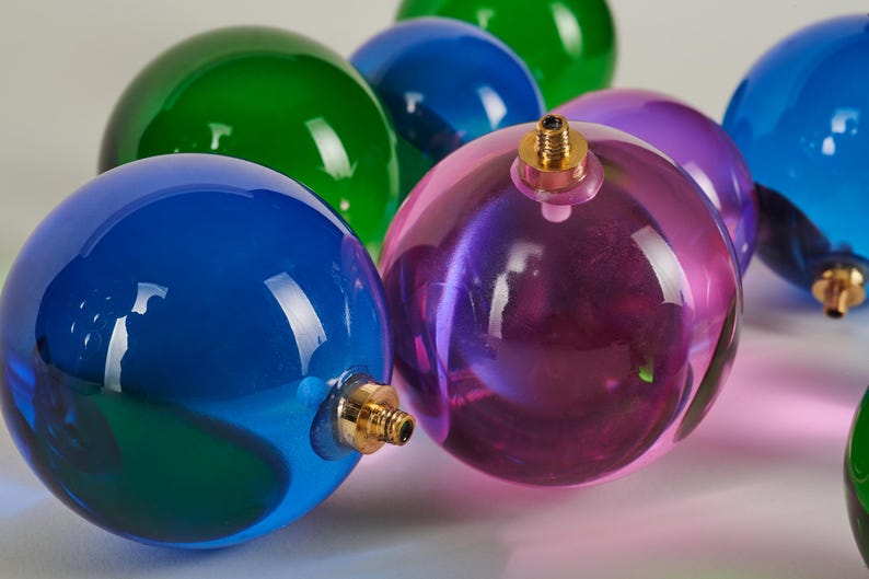 May include: A collection of colorful, transparent glass ornaments in shades of blue, green, and purple. Each sphere has a gold-colored metal cap. The ornaments are arranged closely together, reflecting light and creating a festive display.