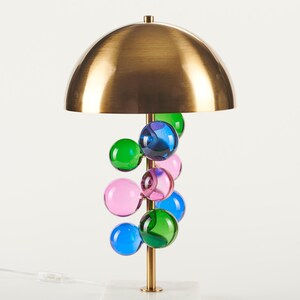 May include: A modern table lamp with a brass dome shade and a cluster of colorful glass orbs. The lamp has a brass stem and a white marble base. The glass orbs are in shades of blue, green, and pink.