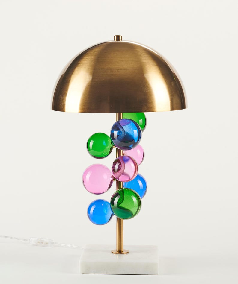 May include: A modern table lamp with a brass dome shade and a cluster of colorful glass orbs. The lamp has a brass stem and a white marble base. The glass orbs are in shades of blue, green, and pink.
