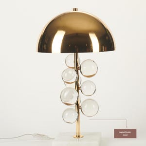 May include: A gold-finished table lamp with a domed shade and a stem adorned with clear glass spheres. The lamp sits on a white marble base. The metal finish is gold.