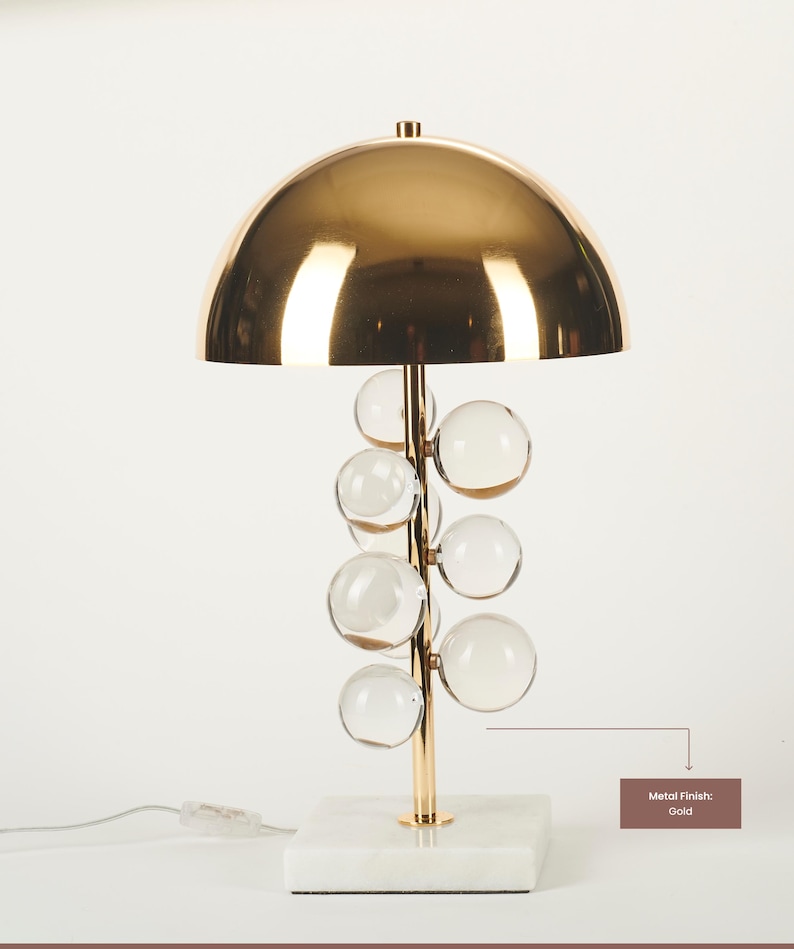 May include: A gold-finished table lamp with a domed shade and a stem adorned with clear glass spheres. The lamp sits on a white marble base. The metal finish is gold.