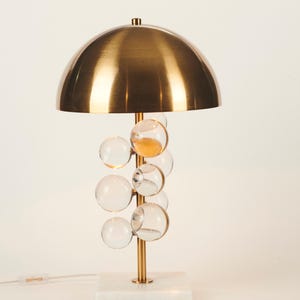 May include: A modern table lamp with a brass dome shade and a cluster of clear glass bubble accents. The lamp has a gold-colored stem and sits on a white marble base. The lamp is approximately 18 inches tall.