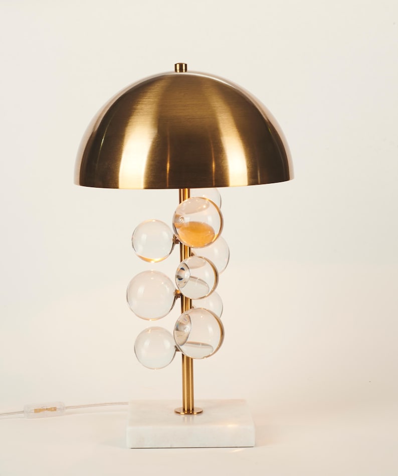 May include: A modern table lamp with a brass dome shade and a cluster of clear glass bubble accents. The lamp has a gold-colored stem and sits on a white marble base. The lamp is approximately 18 inches tall.