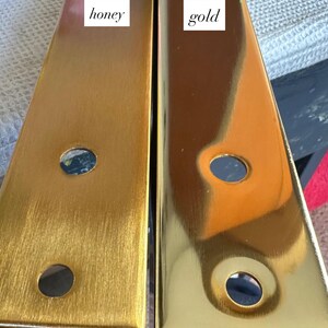 May include: Two rectangular metal furniture legs, one with a honey finish and the other with a gold finish. Each leg has two circular holes. The honey leg has a brushed texture, while the gold leg is highly polished. The word "honey" and "gold" are written on the legs.