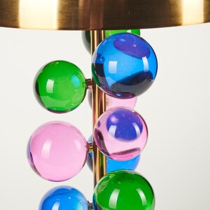 May include: A modern table lamp with a brass finish and a series of colorful glass spheres in green, blue, and pink. The lamp has a large, gold-colored shade.
