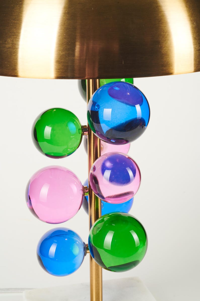 May include: A modern table lamp with a brass finish and a series of colorful glass spheres in green, blue, and pink. The lamp has a large, gold-colored shade.