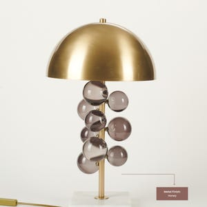 May include: A modern table lamp with a honey-colored metal dome shade and a cluster of smoky glass spheres. The lamp has a gold-colored stem and sits on a white marble base. The text on the image says "Metal Finish: Honey".