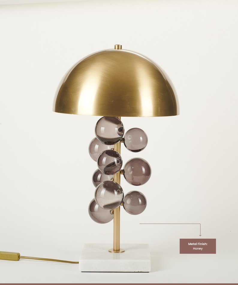 May include: A modern table lamp with a honey-colored metal dome shade and a cluster of smoky glass spheres. The lamp has a gold-colored stem and sits on a white marble base. The text on the image says "Metal Finish: Honey".