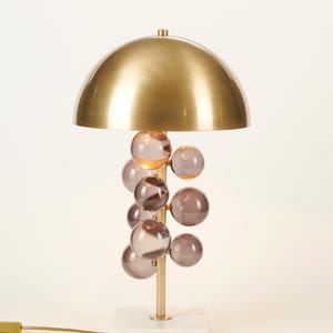 May include: A modern table lamp with a gold-colored, dome-shaped shade. The lamp features a cluster of smoky-purple glass orbs and a gold-colored stem, set on a white marble base. The lamp has a cord with a plug.