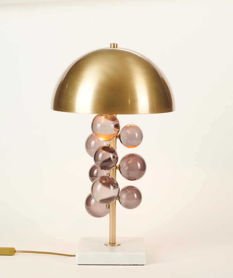 May include: A modern table lamp with a gold-colored, dome-shaped shade. The lamp features a cluster of smoky-purple glass orbs and a gold-colored stem, set on a white marble base. The lamp has a cord with a plug.