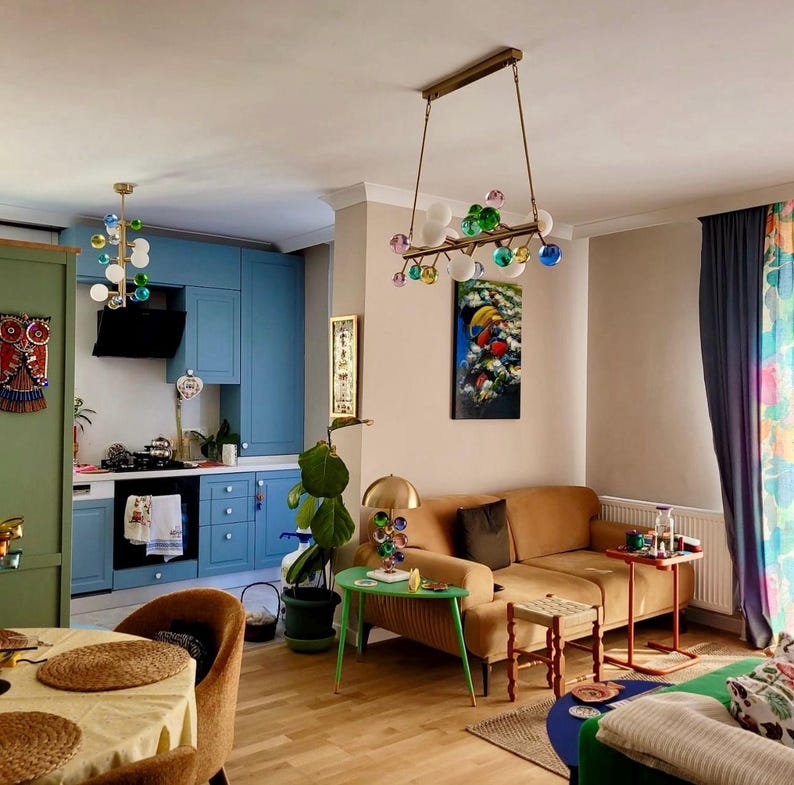 May include: A living room with a modern design, featuring a tan sofa, a green side table, and a colorful painting. Two unique light fixtures with glass globes hang from the ceiling. The kitchen area has blue cabinets and a black range hood.