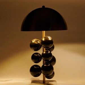 May include: A modern table lamp with a black dome shade and a brass-colored stem. The lamp features a series of black spherical accents and sits on a square, marble-like base. The design is sleek and contemporary.