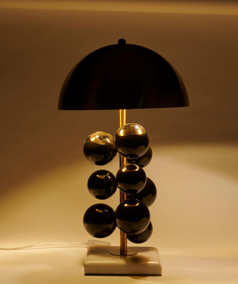 May include: A modern table lamp with a black dome shade and a brass-colored stem. The lamp features a series of black spherical accents and sits on a square, marble-like base. The design is sleek and contemporary.