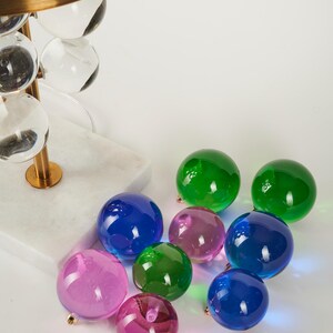 May include: A modern lamp with a brass shade and clear glass orb accents. Scattered around the base are colorful glass spheres in shades of green, blue, and pink. The lamp base is white marble.