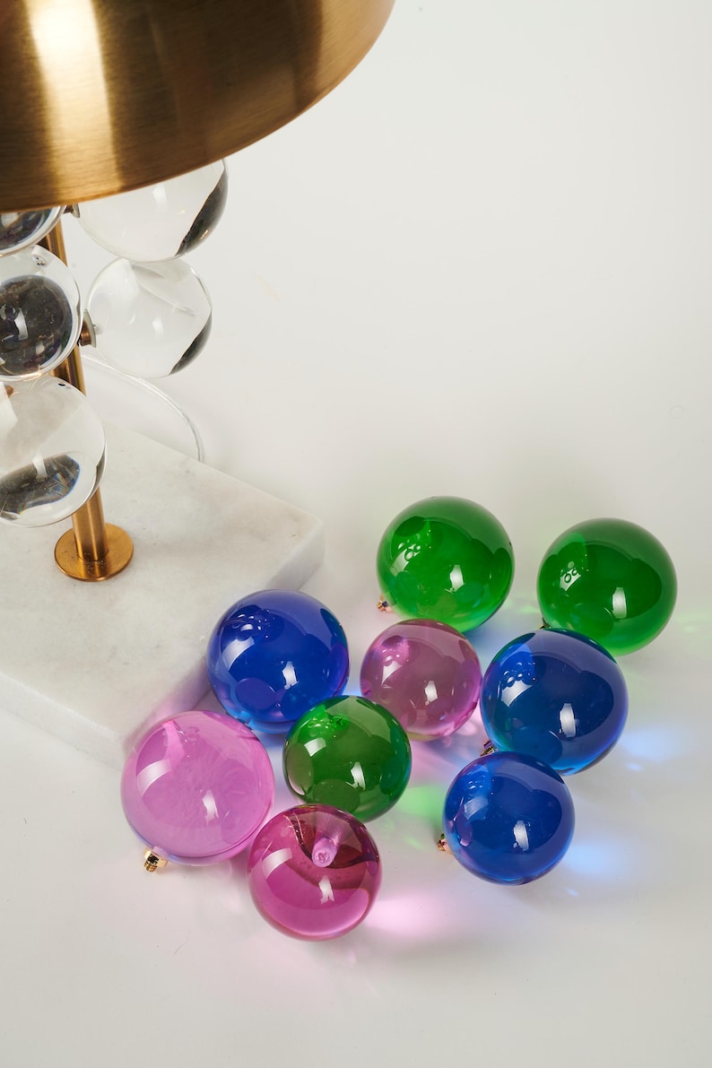 May include: A modern lamp with a brass shade and clear glass orb accents. Scattered around the base are colorful glass spheres in shades of green, blue, and pink. The lamp base is white marble.