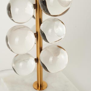 May include: A modern table lamp with a gold-colored metal frame and a white marble base. The lamp features clear glass spheres arranged vertically, and a gold-colored, semi-circular shade.