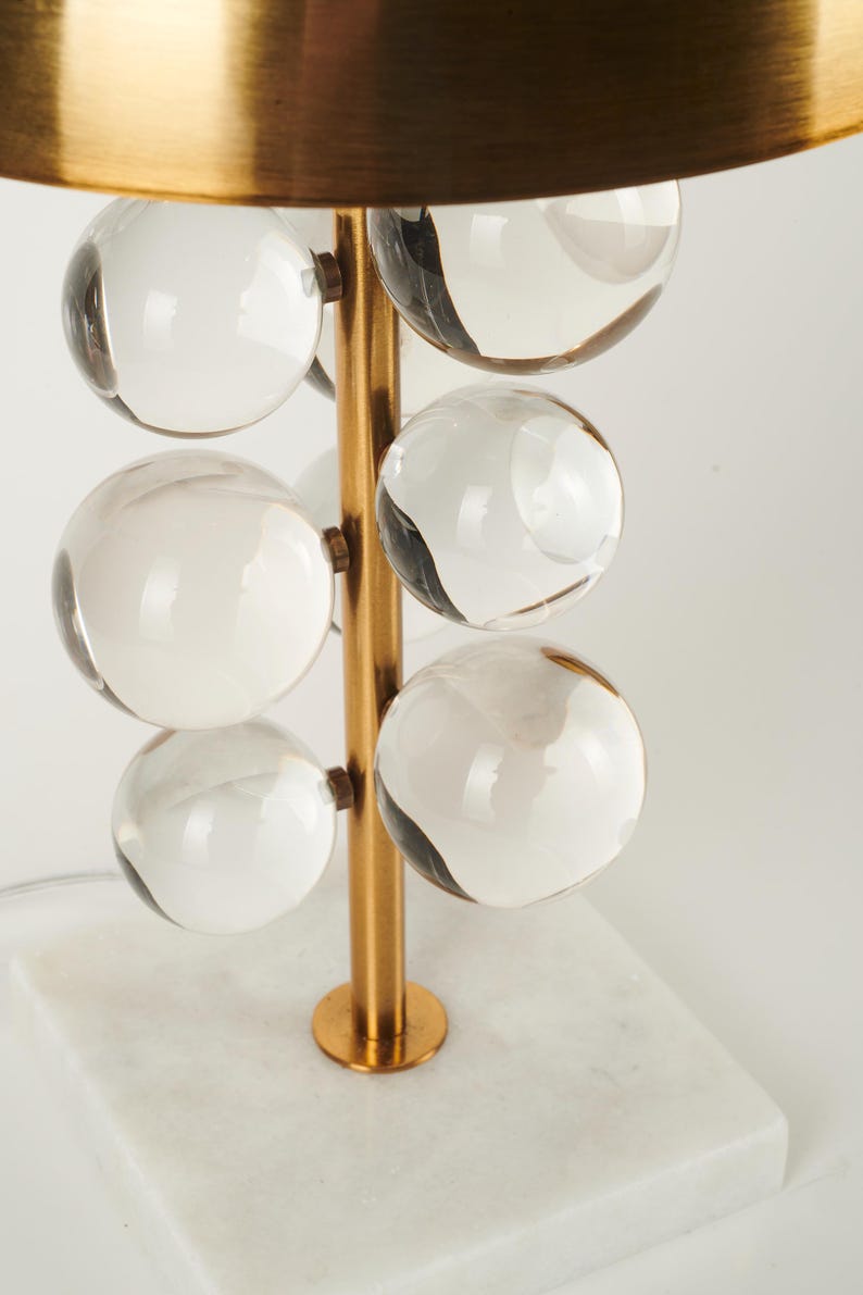 May include: A modern table lamp with a gold-colored metal frame and a white marble base. The lamp features clear glass spheres arranged vertically, and a gold-colored, semi-circular shade.