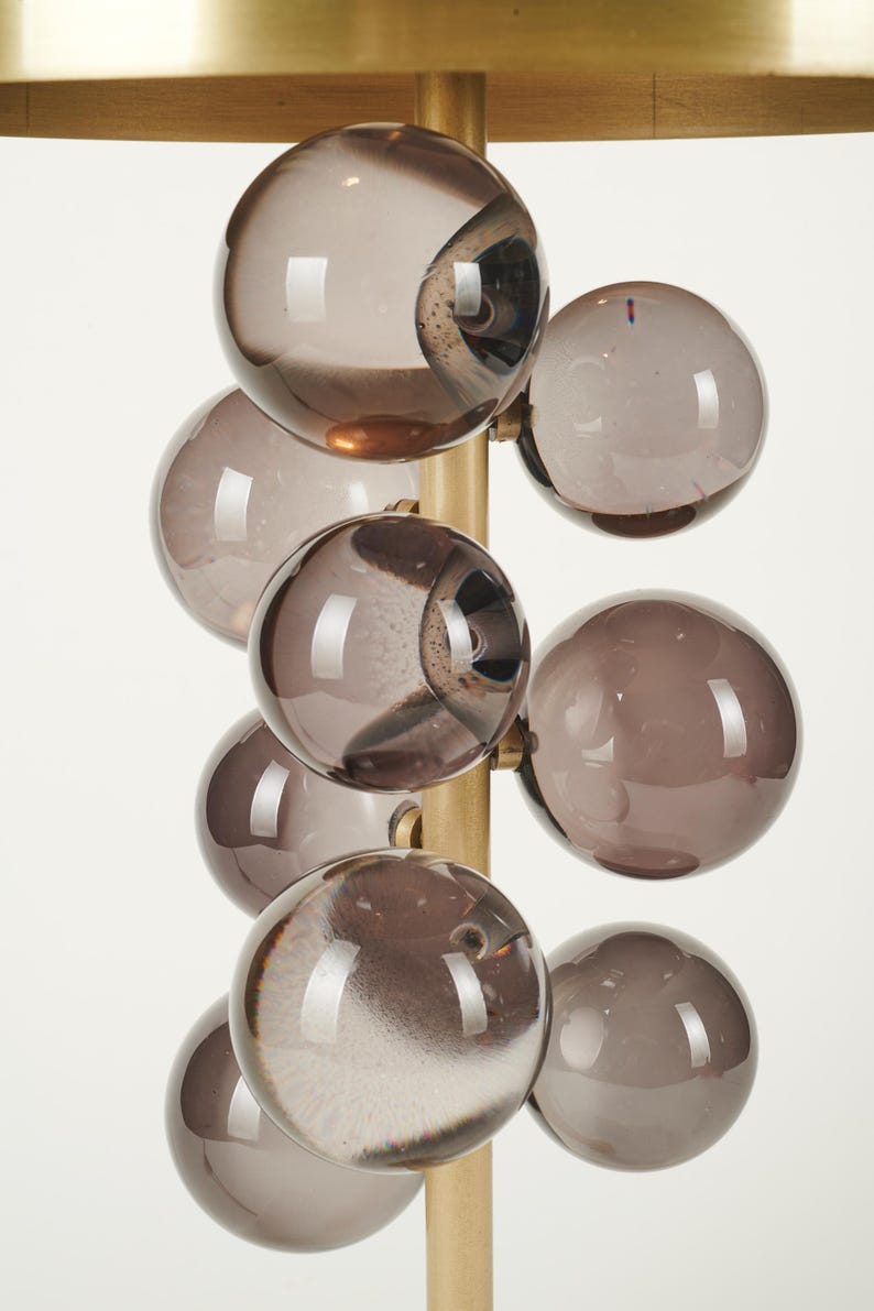 May include: A modern lamp featuring a cluster of smoky gray glass globes attached to a brass-colored metal frame. The lamp has a gold-colored, rectangular top. The glass globes are arranged in a cascading pattern.