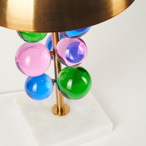 May include: A modern table lamp with a brass-colored stem and a gold-colored shade. The lamp features colorful glass spheres in pink, blue, and green. The base is a white marble square.