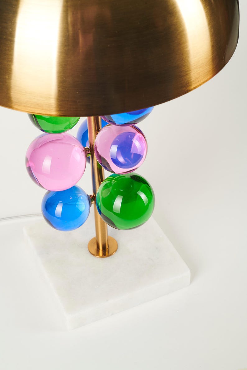 May include: A modern table lamp with a brass-colored stem and a gold-colored shade. The lamp features colorful glass spheres in pink, blue, and green. The base is a white marble square.