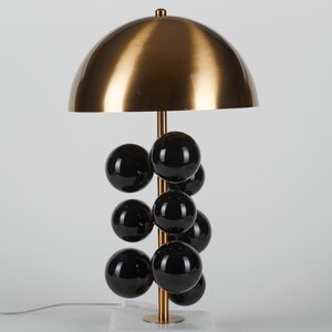 May include: A modern table lamp with a brass-colored dome shade and a vertical stem adorned with black spherical accents. The lamp rests on a white marble base, and has a brass-colored stem. The lamp is a stylish lighting fixture.