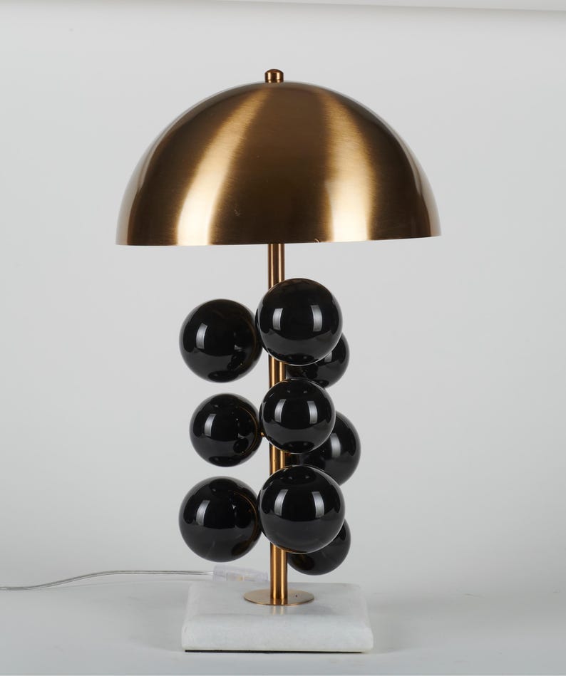 May include: A modern table lamp with a brass-colored dome shade and a vertical stem adorned with black spherical accents. The lamp rests on a white marble base, and has a brass-colored stem. The lamp is a stylish lighting fixture.