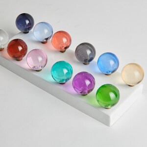 May include: A collection of twelve colorful glass knobs in various shades, including clear, red, pink, green, and blue. Each knob is round and mounted on a white rectangular base, showcasing a range of decorative hardware options.