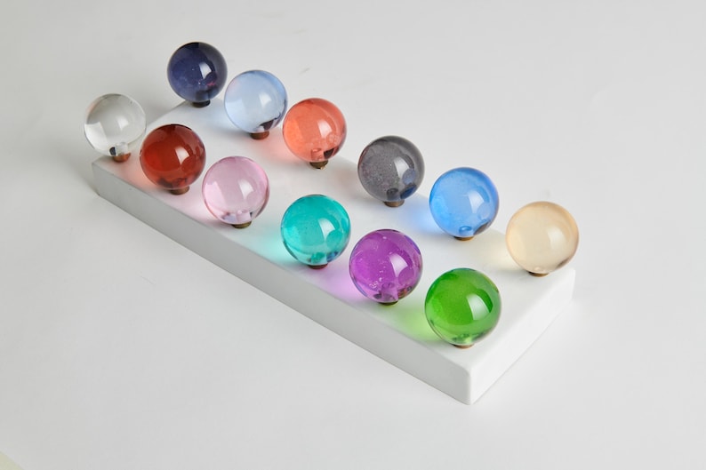 May include: A collection of twelve colorful glass knobs in various shades, including clear, red, pink, green, and blue. Each knob is round and mounted on a white rectangular base, showcasing a range of decorative hardware options.