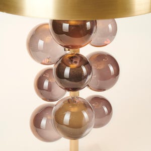 May include: A modern table lamp featuring a brass shade and a stem adorned with multiple brown glass spheres. The lamp has a gold base and a white marble base. The design is contemporary and decorative.