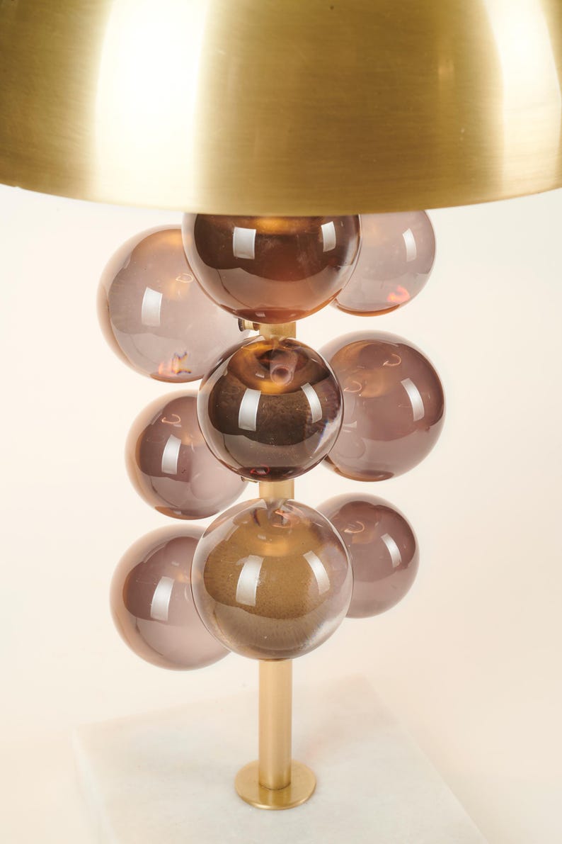 May include: A modern table lamp featuring a brass shade and a stem adorned with multiple brown glass spheres. The lamp has a gold base and a white marble base. The design is contemporary and decorative.