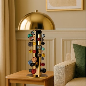 May include: A gold-colored table lamp with a domed shade and a cylindrical base. The base is adorned with a cascade of colorful glass spheres in various sizes and hues, including red, blue, and yellow. The lamp sits on a wooden side table.