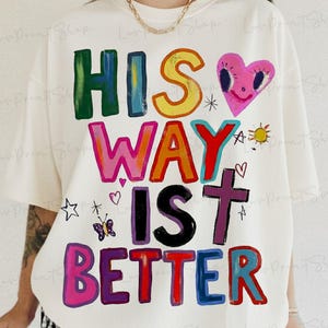 May include: Cream-colored t-shirt with the phrase "HIS WAY IS BETTER" in colorful, block letters. The design includes a pink heart, a cross, a sun, and other small illustrations. The shirt is a casual, oversized fit.