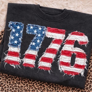 Puede incluir: Sudadera gris oscuro con el número "1776" aplicado en la parte delantera. Los números están diseñados para parecerse a la bandera estadounidense, con campos de estrellas azules y rayas rojas y blancas.