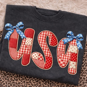 May include: Dark gray sweatshirt with the word "USA" in red, white, and blue patchwork letters. Each letter has a different pattern, including gingham and polka dots. Blue bows with white stars accent the letters. The sweatshirt is displayed on a leopard print surface.
