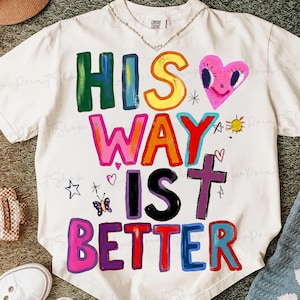 Puede incluir: Camiseta de color crema con la frase "HIS WAY IS BETTER" en letras de bloque coloridas. El diseño incluye un corazón rosa, una cruz, una mariposa y otras pequeñas ilustraciones. La camiseta tiene mangas cortas y cuello redondo.