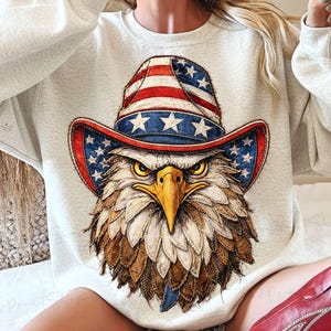 以下が含まれることがあります： 愛国的なカウボーイハットをかぶったワシの精巧なイラストが描かれた、明るい灰色のスウェットシャツ。帽子は赤と白のストライプ、青い星、白い星で装飾されています。ワシは厳しい表情をしています。