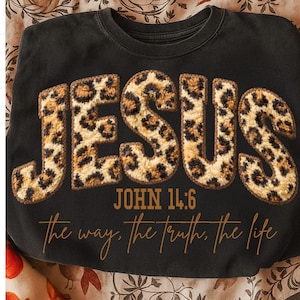 May include: Black sweatshirt featuring the word "JESUS" in leopard print lettering. The text "JOHN 14:6" and the phrase "the way, the truth, the life" are below the word "JESUS" in a script font.