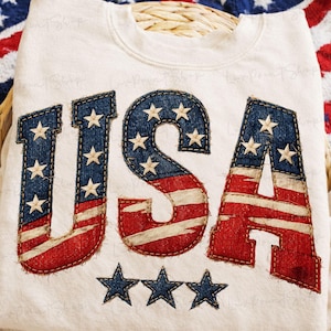 May include: Cream-colored sweatshirt with the word "USA" appliqued in denim and red fabric, featuring stars and stripes. The letters are outlined with a contrasting stitch, and three blue stars are below.