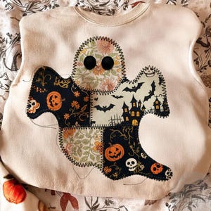 Op de afbeelding: Crèmekleurige sweatshirt met een patchwork spookontwerp. De geest heeft bloemen-, pompoen- en spookhuispatronen. Zwarte knoopogen en een gestikte omtrek maken de Halloween-thema kleding compleet.