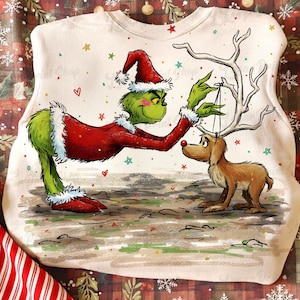 Könnte beinhalten: Weißes Sweatshirt mit einer Cartoon-Illustration des Grinch in einem Weihnachtsmann-Anzug, der einem Rentier ein Geweih aufsetzt. Der Grinch ist grün mit rotem Weihnachtsmannhut und -anzug. Das Rentier ist braun. Sterne und Herzen sind verstreut.