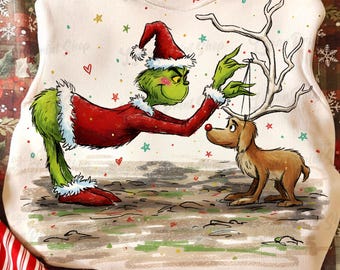 May include: A white sweatshirt featuring a cartoon illustration of the Grinch in a Santa suit, placing antlers on a reindeer. The Grinch is green with a red Santa hat and suit. The reindeer is brown. Stars and hearts are scattered around the image.