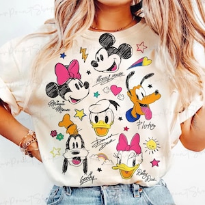 Puede incluir: Camiseta color crema con ilustraciones de dibujos animados de Mickey Mouse, Minnie Mouse, Donald Duck, Daisy Duck, Goofy y Pluto. El dise&ntilde;o incluye estrellas, corazones y los nombres de los personajes. Una camiseta gr&aacute;fica divertida y colorida.