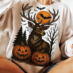May include: A cream-colored sweatshirt featuring a Halloween-themed graphic. The design includes a brown deer with large antlers, two jack-o'-lanterns, bats, a full orange moon, and a spider web. The overall theme is a spooky autumn scene.