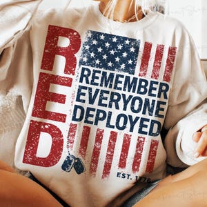 以下が含まれることがあります： クリーム色のスウェットシャツ。色あせたアメリカ国旗のデザインで、赤と青のストライプと星が描かれています。「RED」と「REMEMBER EVERYONE DEPLOYED」の文字は赤と青で表示されています。「EST. 1776」の文字も含まれています。