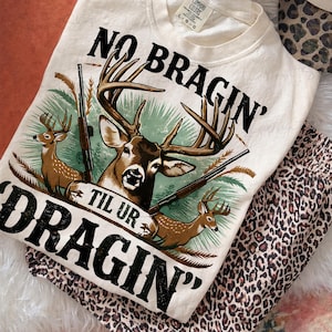 May include: Cream-coloured sweatshirt featuring a deer graphic and the text "NO BRAGIN' TIL UR DRAGIN'". The sweatshirt is paired with leopard print trousers and a matching hat. The design includes deer, rifles, and foliage.
