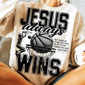 以下が含まれることがあります： バスケットボールをテーマにしたクリーム色のスウェットシャツ。デザインは、黒い文字で「JESUS always WINS」、バスケットボール、バスケットボール選手が特徴です。追加のテキストには聖書の詩が含まれています。