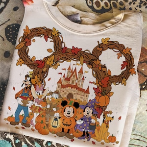 May include: Cream-colored sweatshirt featuring a fall-themed Disney design. The graphic includes Mickey and Minnie Mouse in Halloween costumes, surrounded by pumpkins, autumn leaves, and a castle. The design is framed by a Mickey Mouse-shaped wreath.